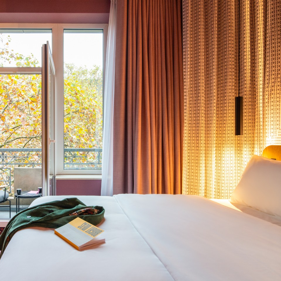 Hotel room with a large bed, warm lighting, a book on the sheets, and an open balcony revealing colorful autumn trees outside.
