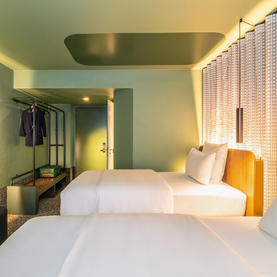 A modern hotel room featuring two twin beds, green walls, stylish lighting, and a minimalist wardrobe with a suit displayed.