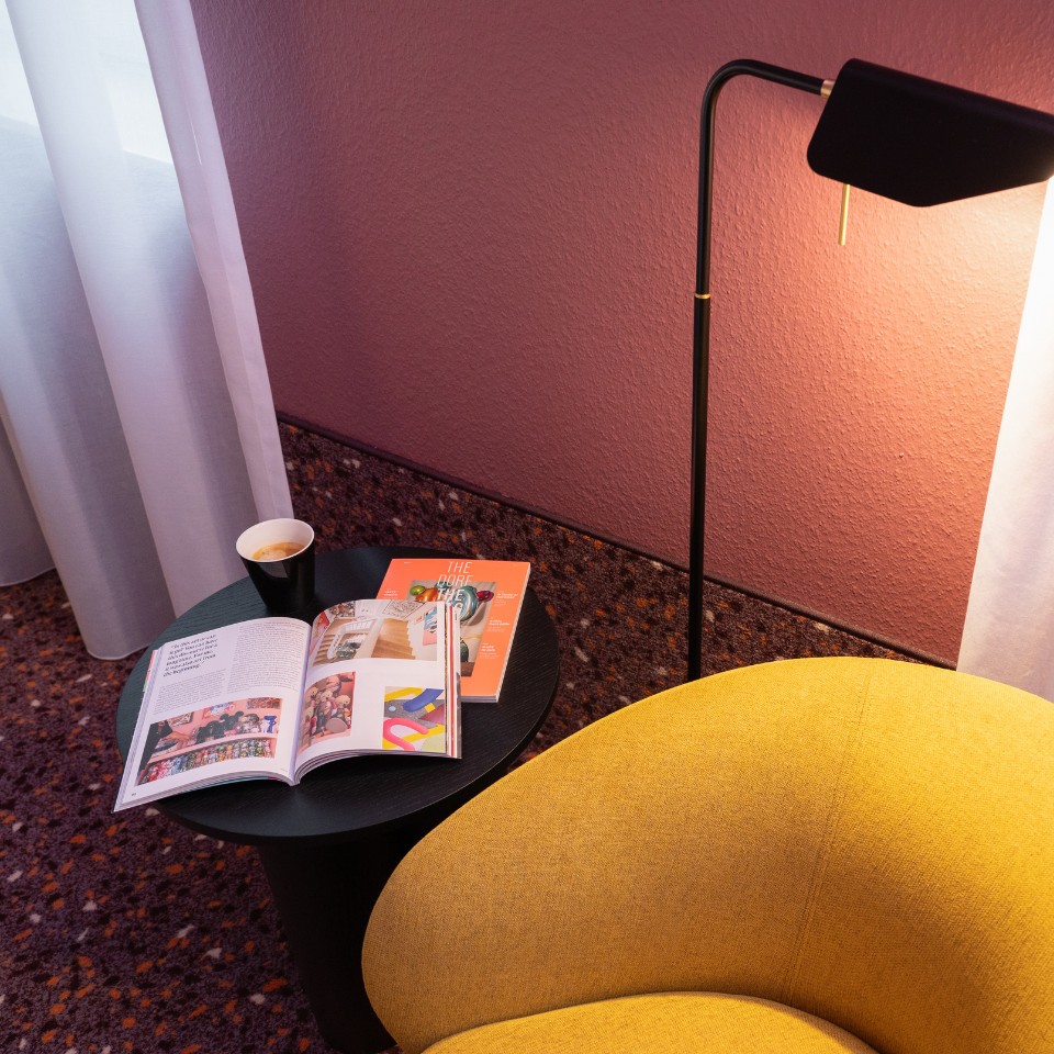 A cozy reading nook featuring a yellow chair, a black side table with magazines and a coffee cup, and a stylish black lamp.