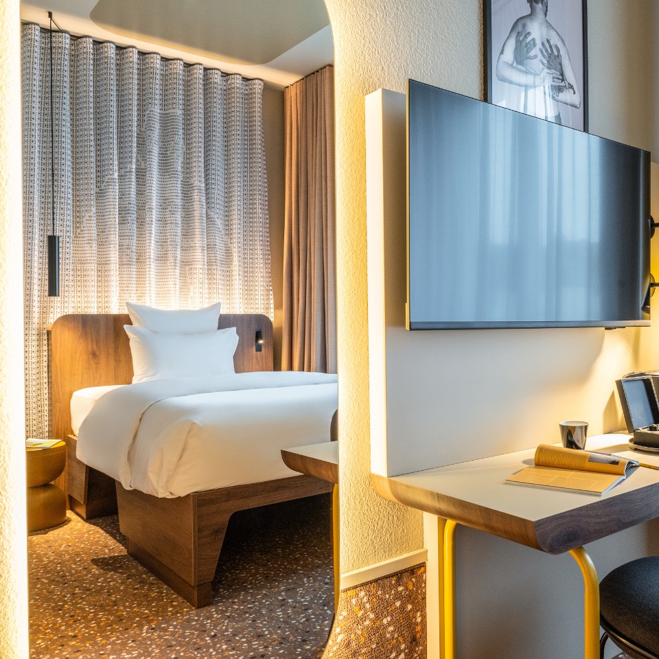 A hotel room featuring a cozy bed, wooden furnishings, a flat-screen TV, and a stylish workspace bathed in natural light.