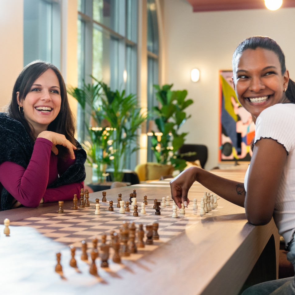 Two individuals engage in a chess game at a stylish venue, surrounded by greenery and modern decor, focused on their strategy.