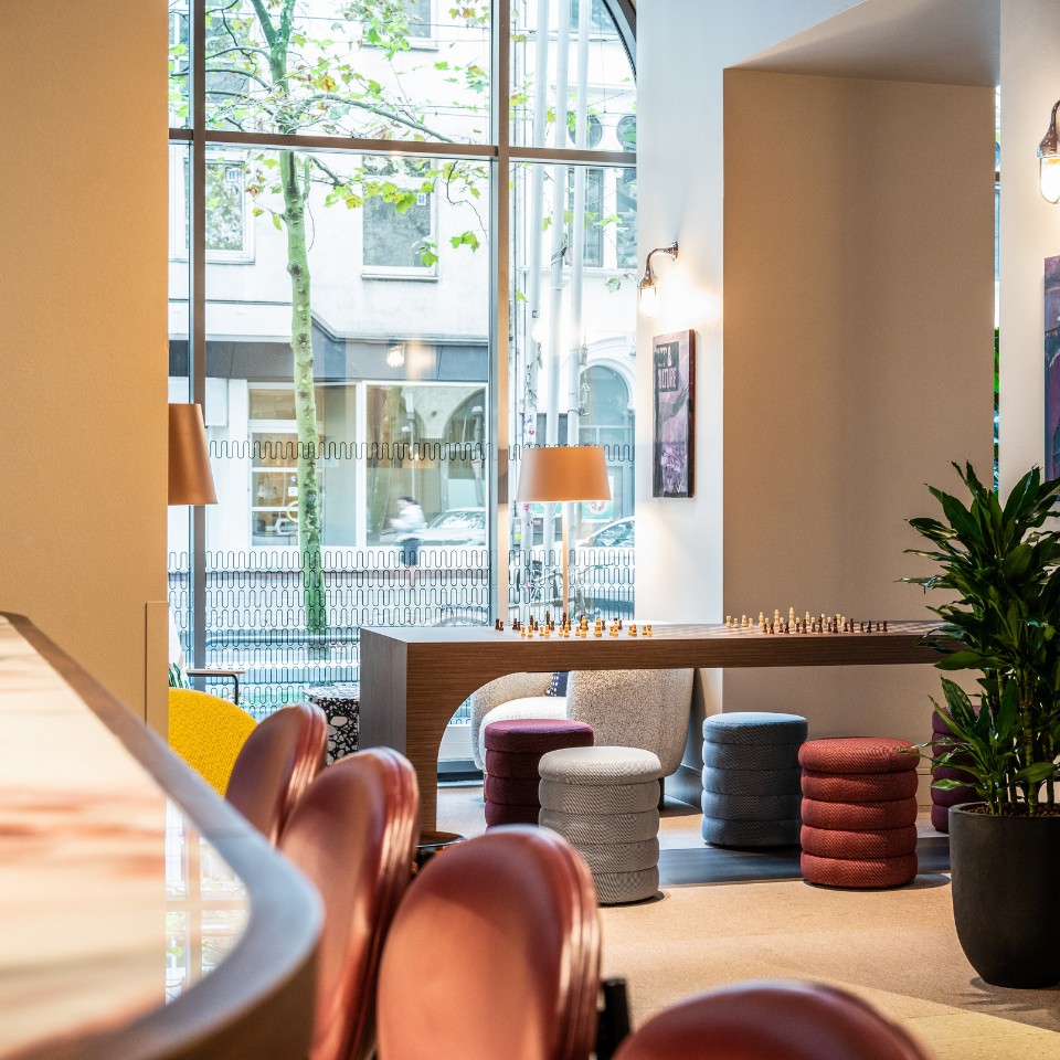 Modern lounge area featuring colorful stools, a chess table, and large windows overlooking a city street, creating a welcoming atmosphere.