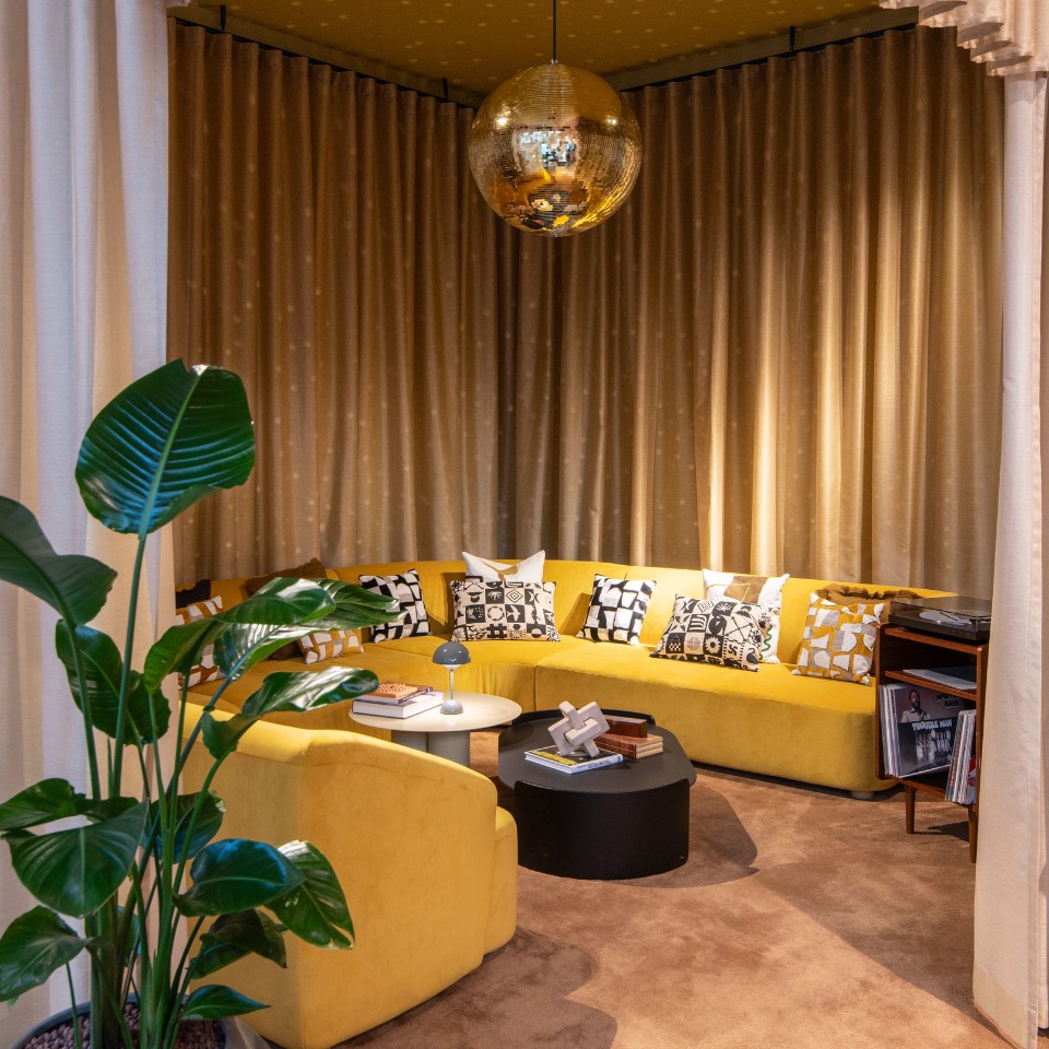 Cozy lounge featuring a yellow sectional sofa adorned with patterned pillows, a disco ball, and potted green plants in warm, ambient lighting.