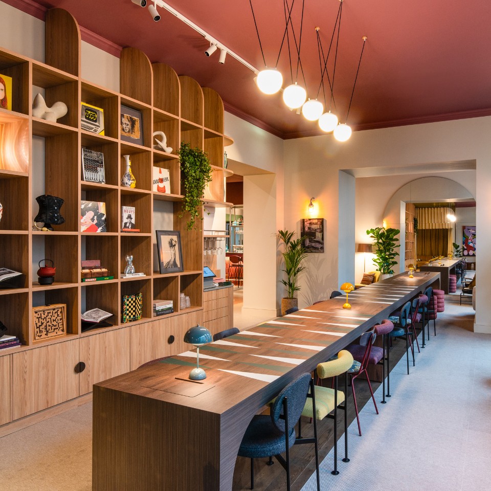 A modern communal space featuring a long table, colorful chairs, shelves with decor, plants, and warm lighting under a pink ceiling.