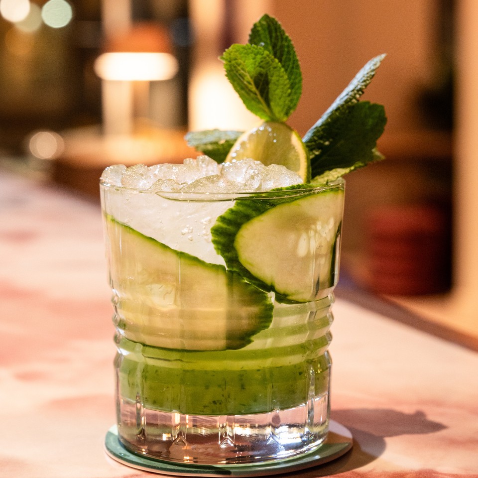 A refreshing cucumber mojito garnished with mint and lime, served in a clear glass filled with ice on a softly lit bar.