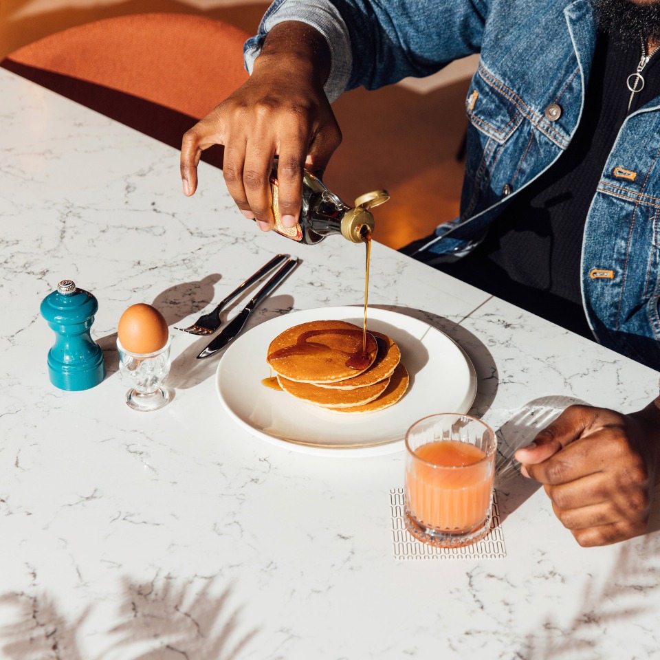 A person pours syrup over fluffy pancakes on a marble table, with an egg, pepper shaker, and juice glass nearby.