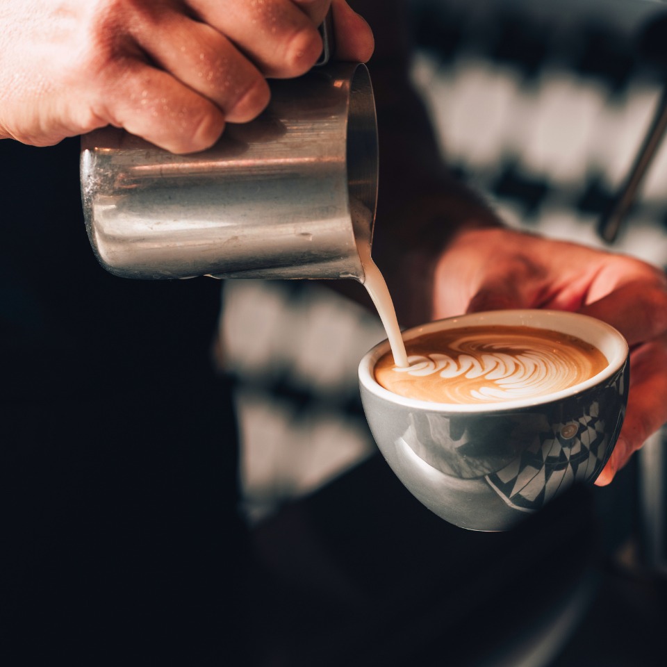 A barista pours steamed milk into a coffee cup, creating intricate latte art on the surface.