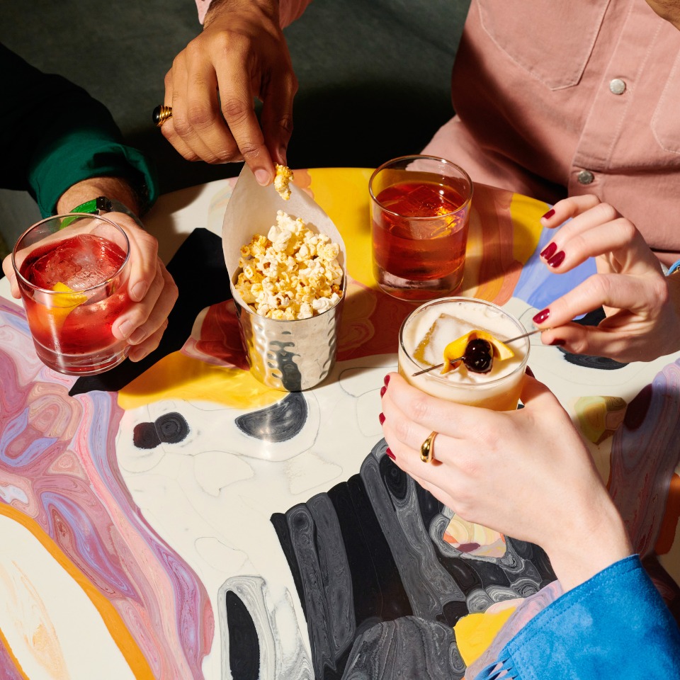 Friends enjoy cocktails and popcorn on a colorful table, with hands reaching for snacks and drinks in a vibrant social setting.