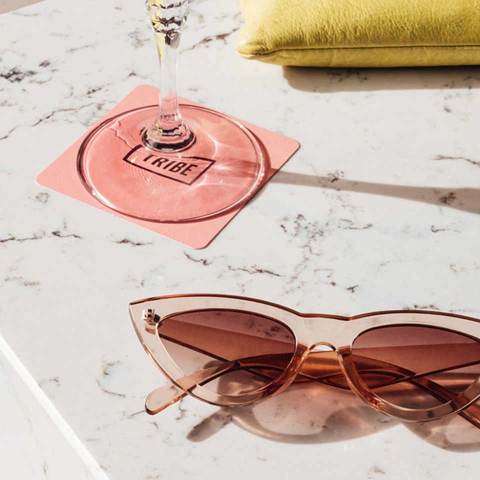 A pair of cat-eye sunglasses rests on a marble table next to a pink coaster and a glass, with a green cushion in the background.