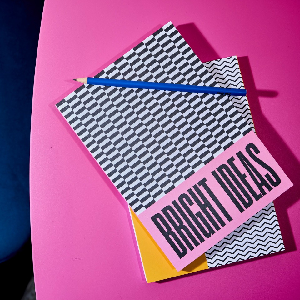 A vibrant pink table features three notebooks: a black and white patterned one, a zigzag design, and a pink one labeled "BRIGHT IDEAS," with a blue pencil.