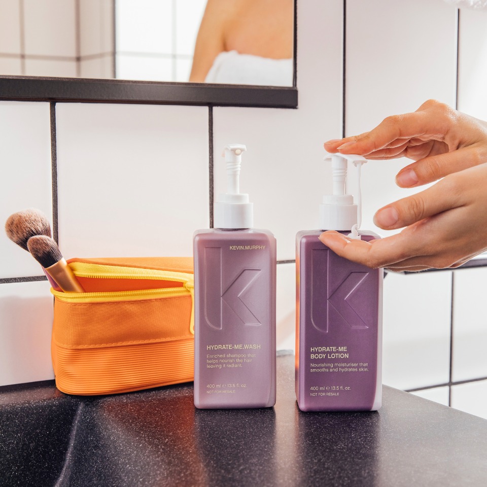 A hand reaches for Kevin Murphy shampoo and body lotion on a bathroom countertop beside a makeup brush and orange pouch.