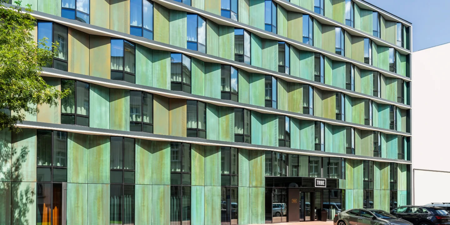 Modern building facade with green metal panels and large windows, featuring the sign "TRIBE" at the entrance. Cars parked nearby.
