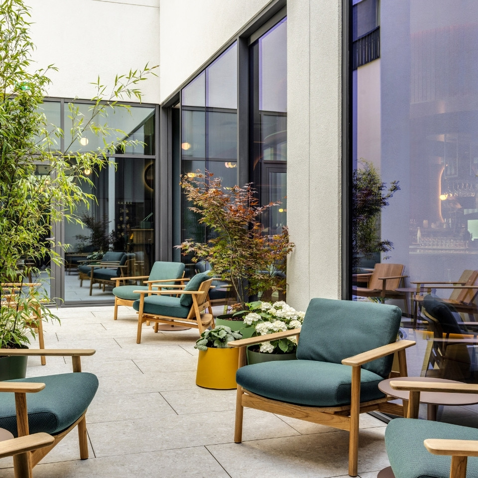 A stylish outdoor lounge area features modern green chairs, potted plants, and large glass windows reflecting a cozy interior.