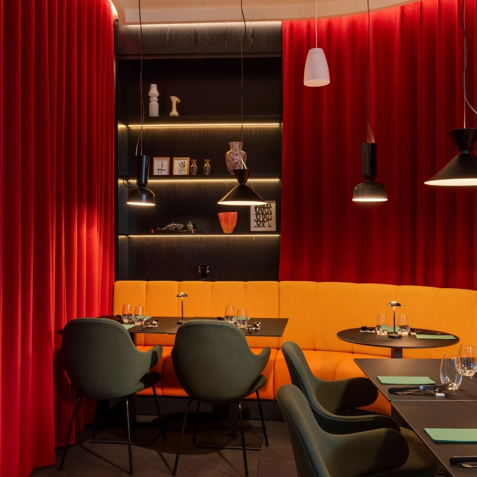Cozy restaurant interior featuring vibrant red curtains, an orange seating area, and elegant black tables with modern lighting.