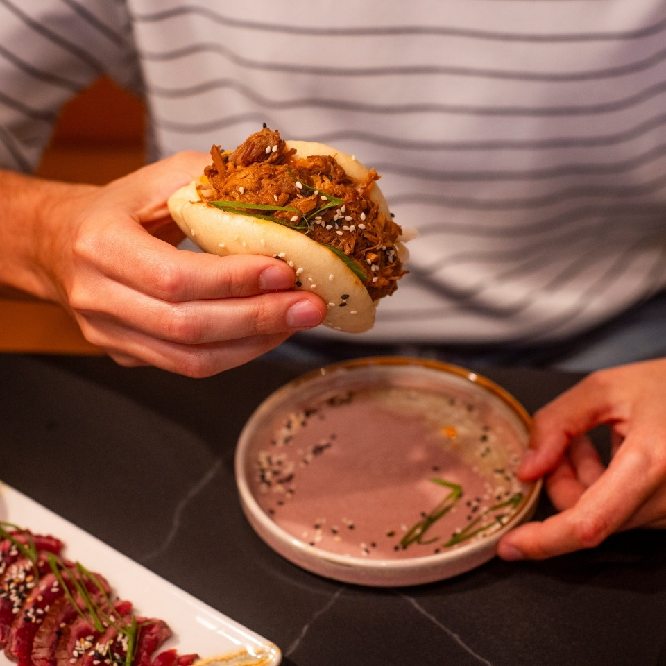 A man holds a sandwich in one hand, with a small bowl of sauce placed beside it on a table.