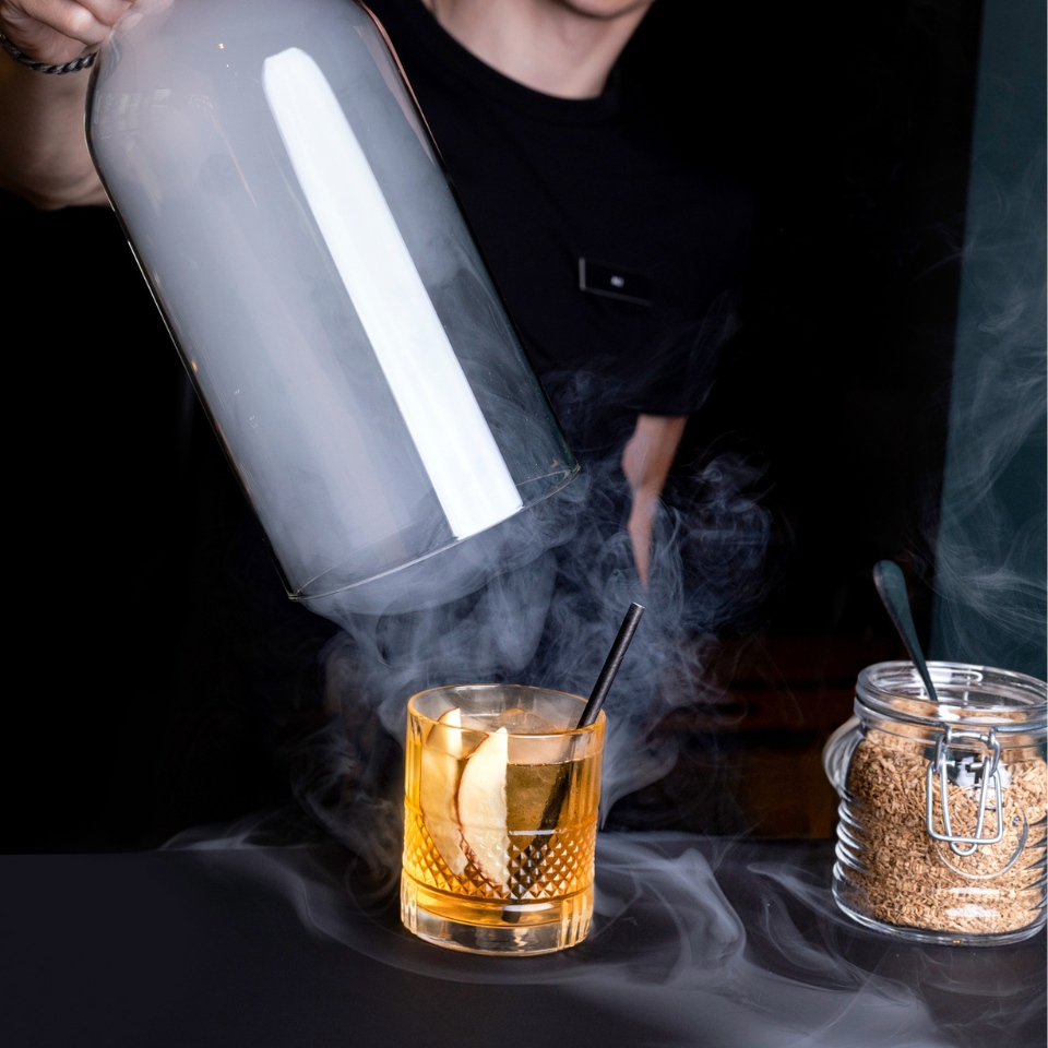 A bartender pouring smoke from a glass dome over a whiskey cocktail garnished with an apple slice, next to a jar of sugar.