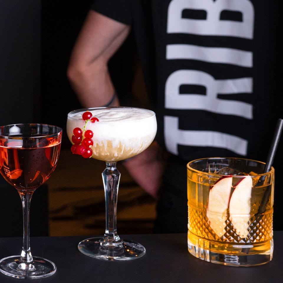 Three cocktails sit on a bar: a red drink, a frothy dessert-like cocktail with berries, and an amber drink with apple slices.
