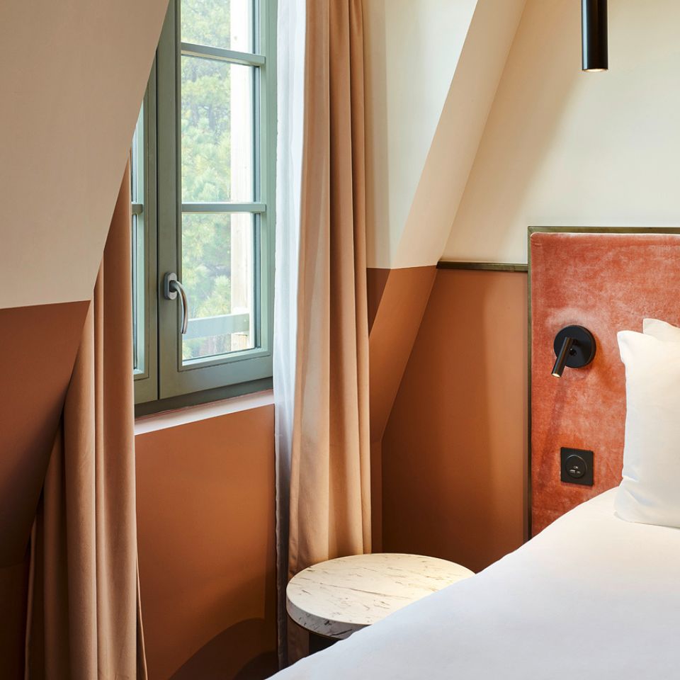 Cozy hotel corner with a green window, soft curtains, and a plush headboard against a warm, earthy-toned wall. A round marble table adds elegance.