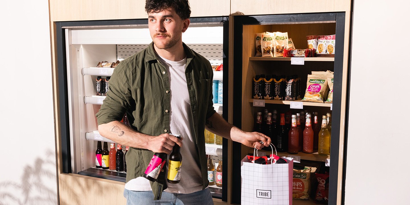 A man stands in front of a fridge, holding two bottles and a shopping bag from TRIBE, surrounded by snacks and drinks.