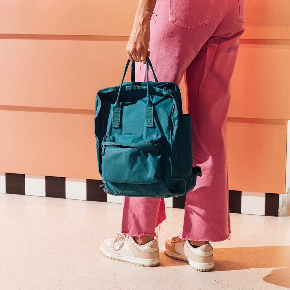 A person wearing pink pants holds a teal backpack in a brightly lit space with an orange wall and checkered floor.