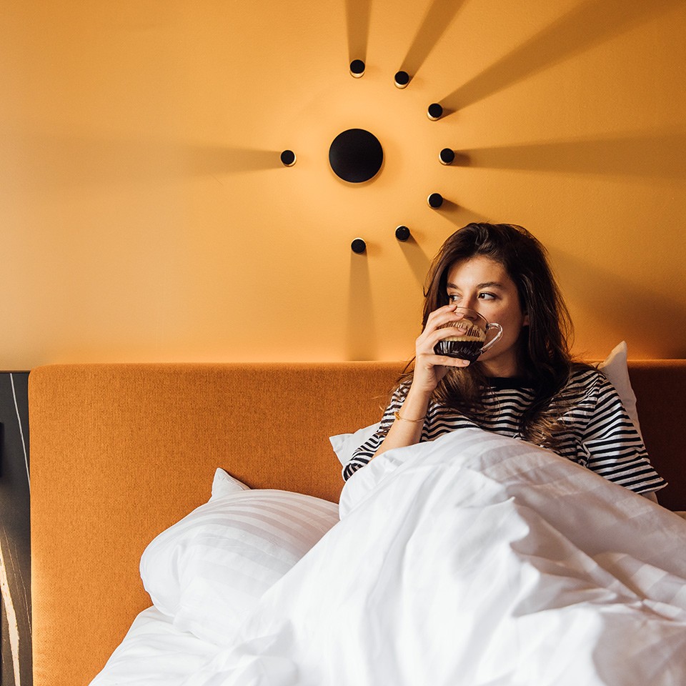 A person sits up in bed, wrapped in white blankets, against a warm orange wall with a stylish black sunburst wall lamp.