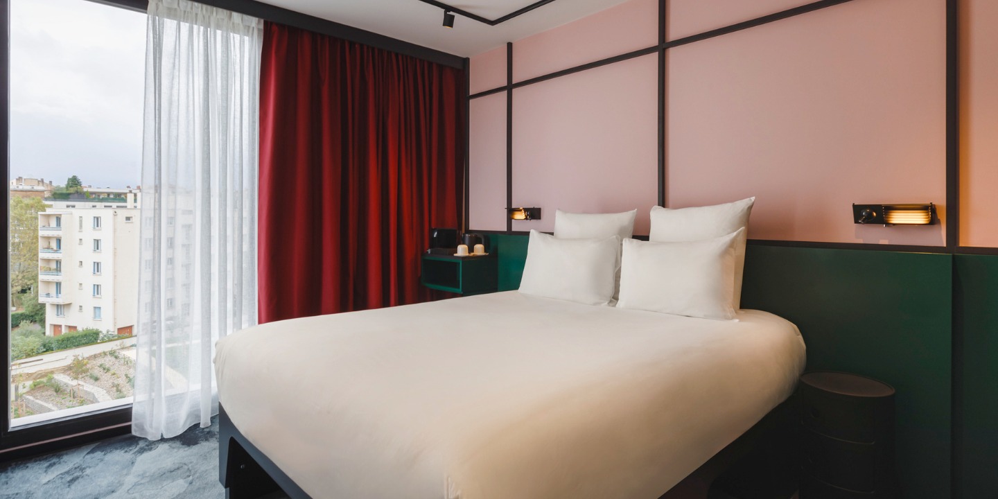 a hotelbed in front of two-toned walls, the view out of the floor-length window shows another building and trees