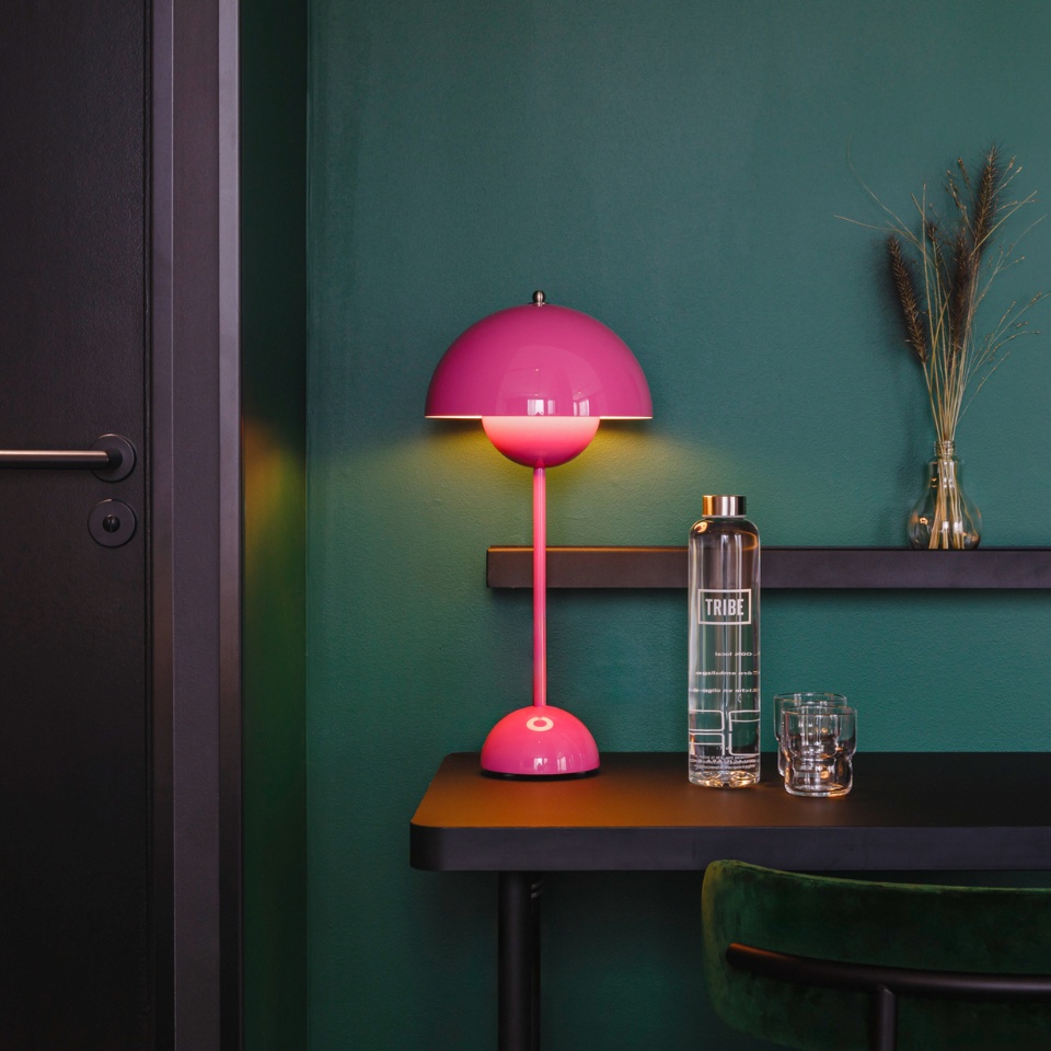 Bright pink lamp illuminates a chic black table with a water bottle, glass, and decorative vase against a deep green wall.