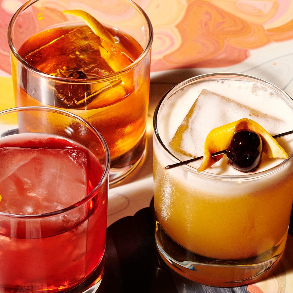 Three stylish cocktails: a red drink with ice, an amber drink with lemon, and a yellow drink garnished with cherry and lemon twist.