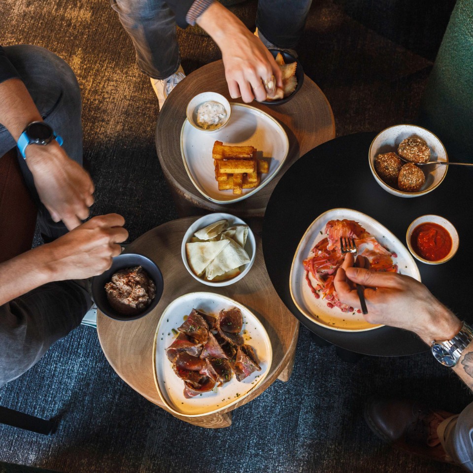 Three people share a variety of dishes on wooden tables, including meats, chips, and dips, creating a cozy dining experience.