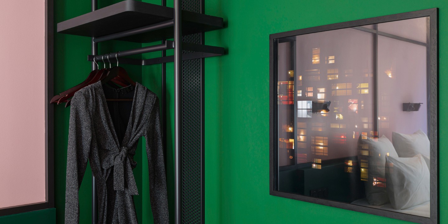 A black dress hangs on a rack against a vibrant green wall, with a painting showing colorful urban lights outside.