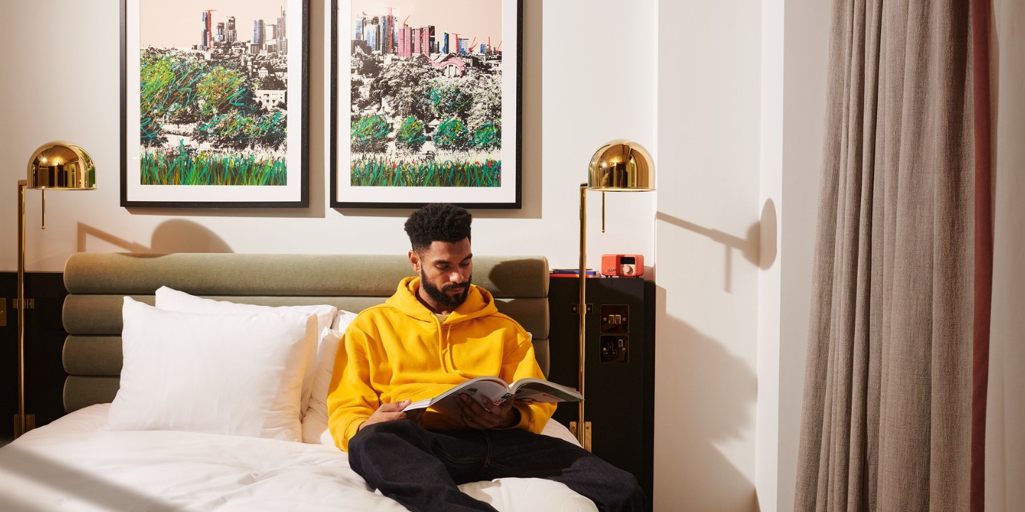 A person in a bright yellow hoodie sits on a bed, reading a magazine with artwork hanging on the wall behind them.