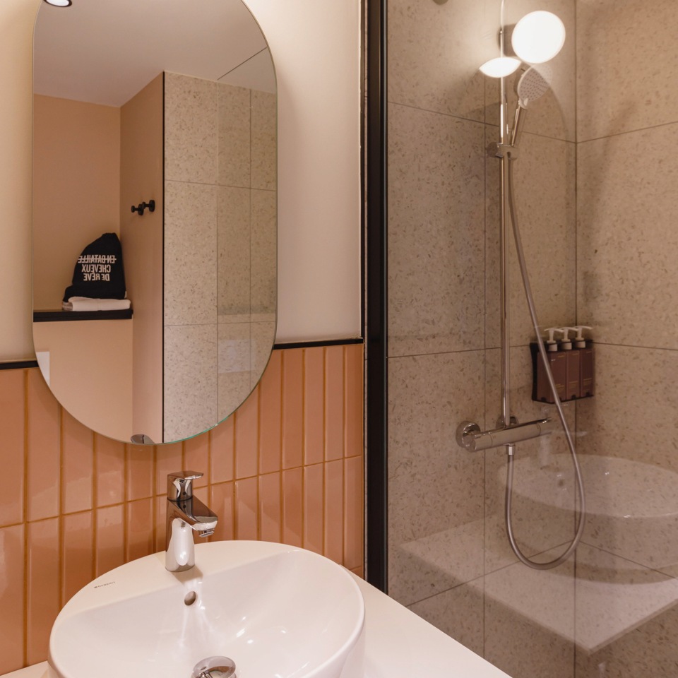 Modern bathroom featuring an oval mirror, a sleek sink, warm orange tiles, and a stylish shower with toiletries on display.