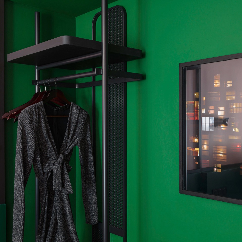 A black dress hangs on a rack against a vibrant green wall, with a painting showing colorful urban lights outside.