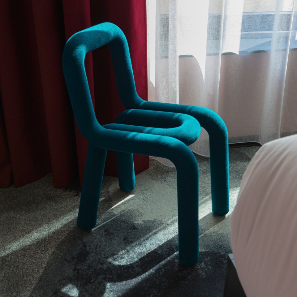 A stylish turquoise chair with rounded edges sits near a window, casting shadows on the floor against red curtains and soft lighting.