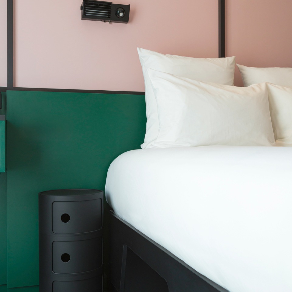 A cozy bedroom corner with a bed featuring white pillows, a dark nightstand, and a green accent wall against a soft pink backdrop.