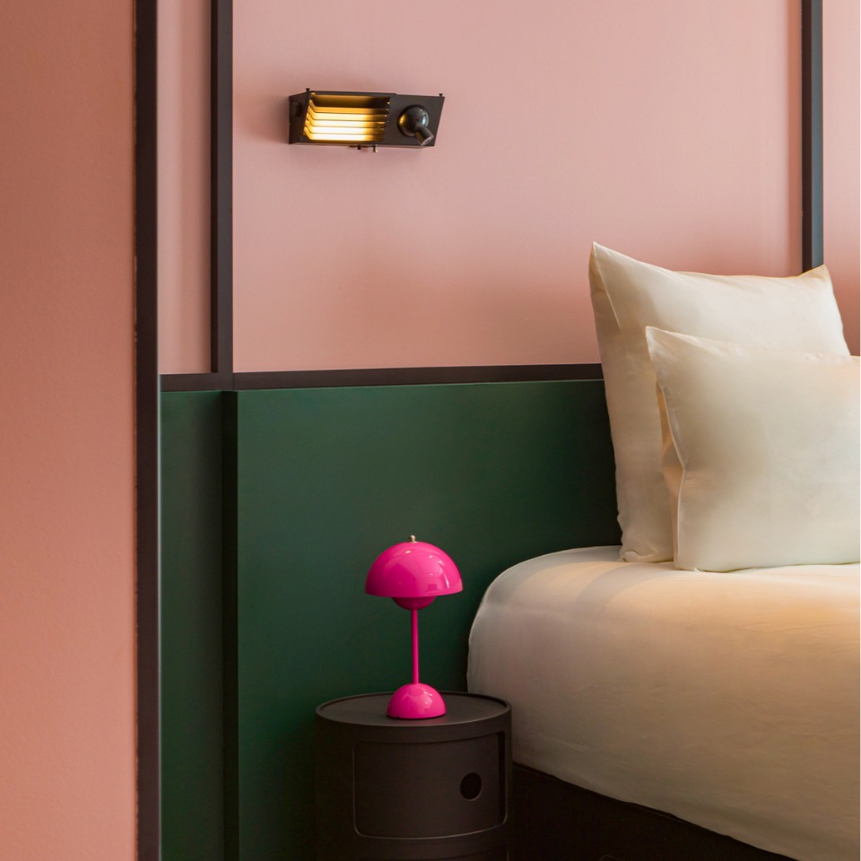 A stylish bedroom corner featuring a pink wall, green accents, plush pillows, and a vibrant pink lamp on a black nightstand.