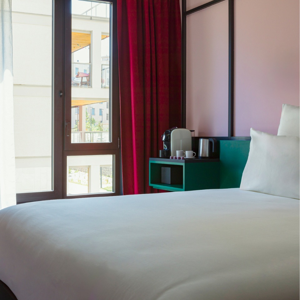 Modern hotel room featuring a white bed, colorful accent furniture, a coffee station, and large windows with red curtains.