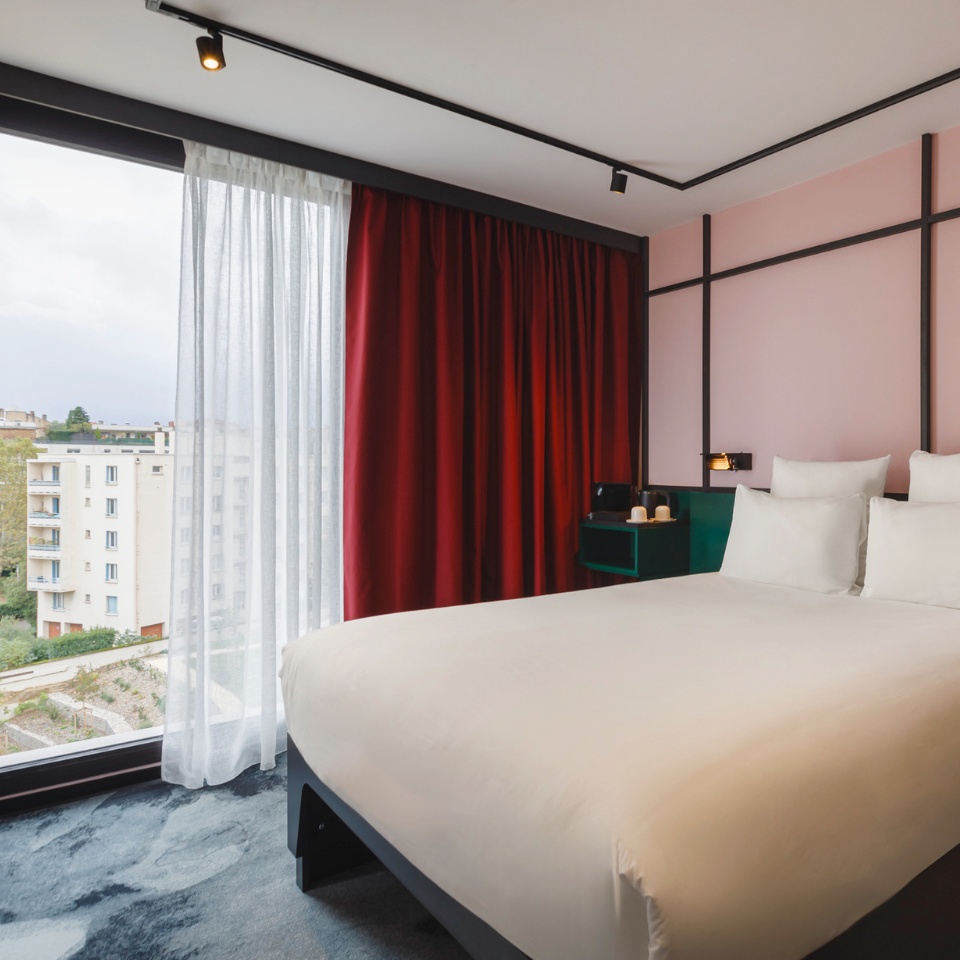 Modern hotel room with a king bed, red curtains, and large windows offering a view of the cityscape outside.