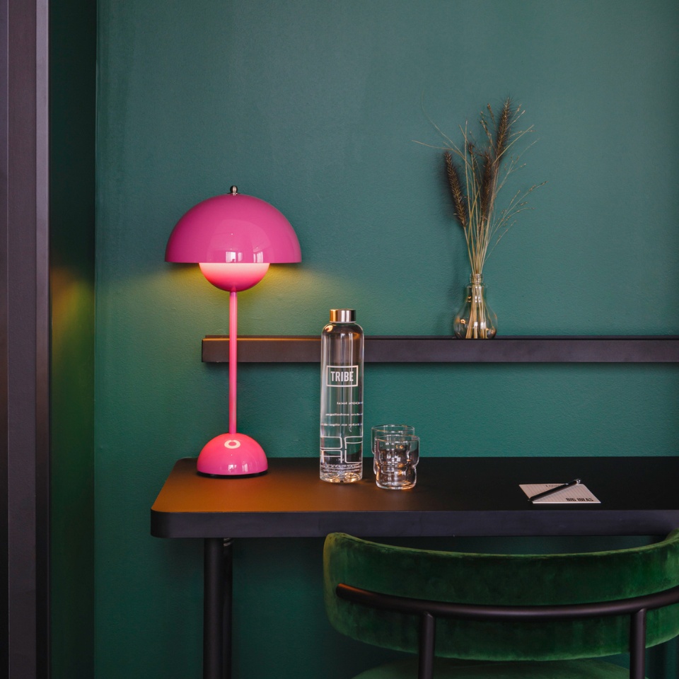 Bright pink lamp illuminates a chic black table with a water bottle, glass, and decorative vase against a deep green wall.