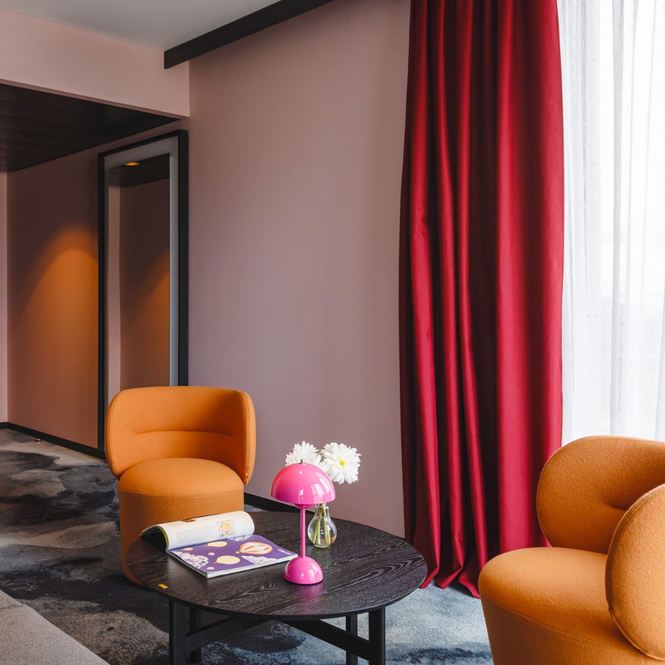 Modern lounge with two orange chairs, a black table, pink lamp, flowers, and red curtains against a soft pink wall.
