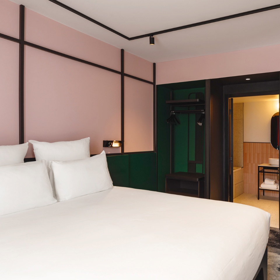 Modern hotel room featuring a king-size bed with white linens, pink and green accent walls, and an attached bathroom.