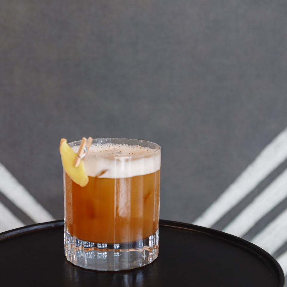 A glass of cocktail with amber liquid, topped with foam and a lemon slice, resting on a black table against a muted backdrop.