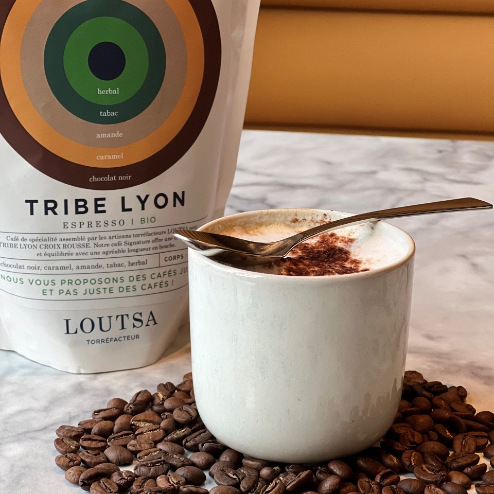 A steaming cup of coffee topped with foam and cocoa lies on a marble surface, surrounded by coffee beans and a bag of Tribe Lyon espresso.
