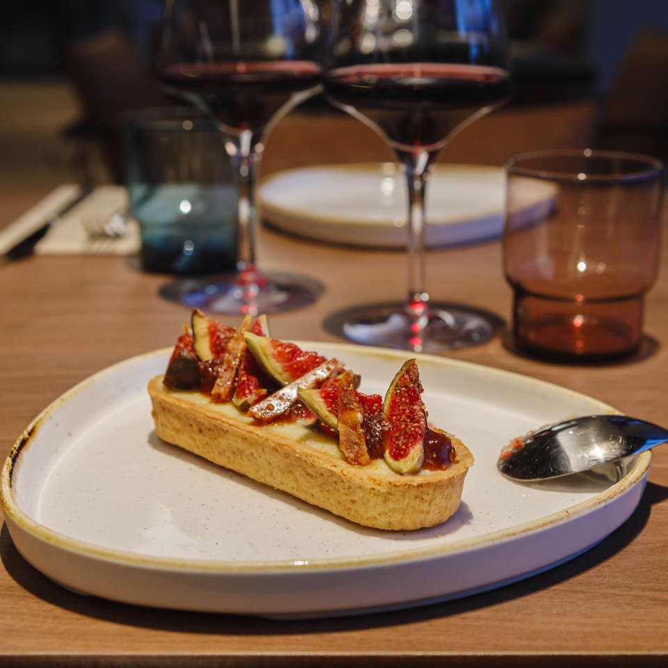 A fig tart topped with fresh figs and caramelized pieces, served on a white plate with red wine glasses in the background.