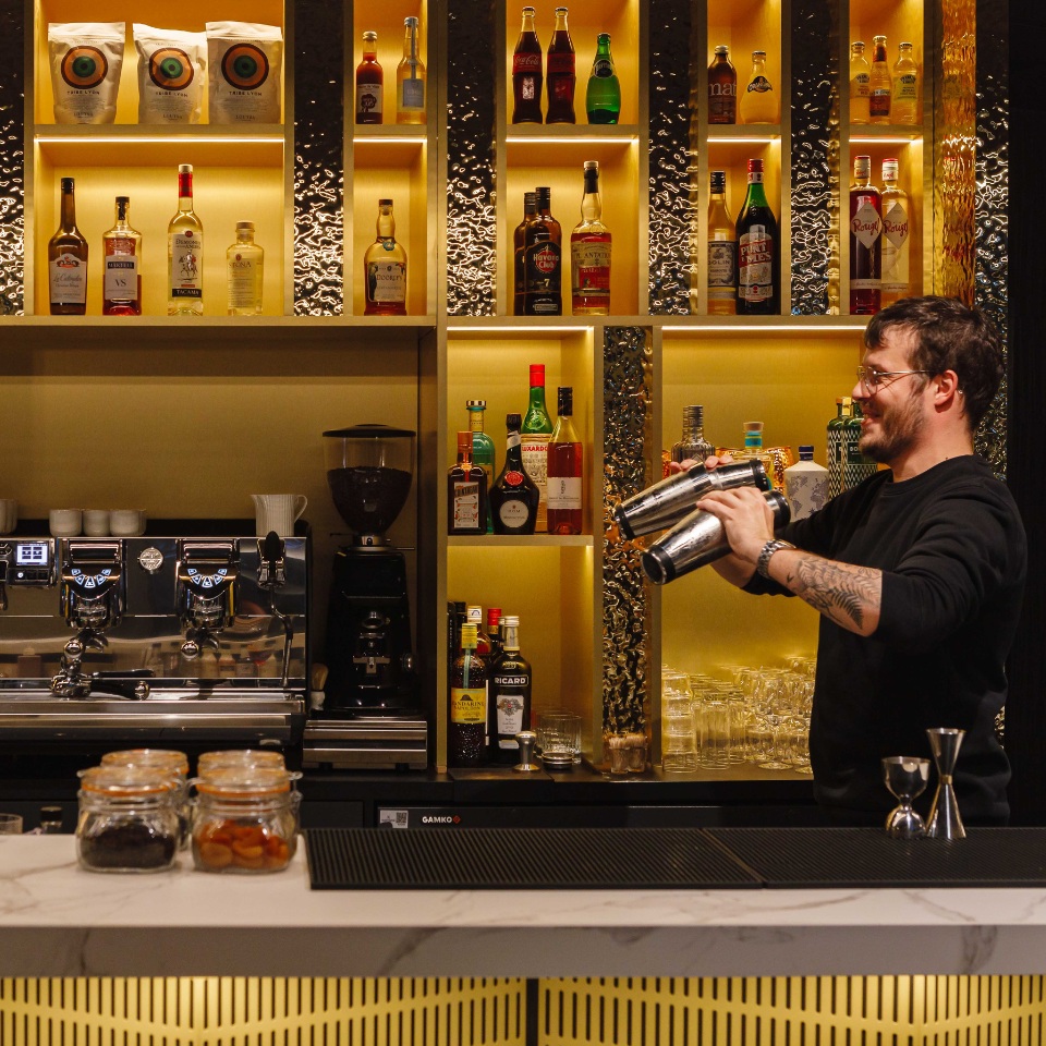 A bartender in a black sweater shakes a cocktail amid shelves stocked with various bottles of spirits and mixers in an elegant bar setup.