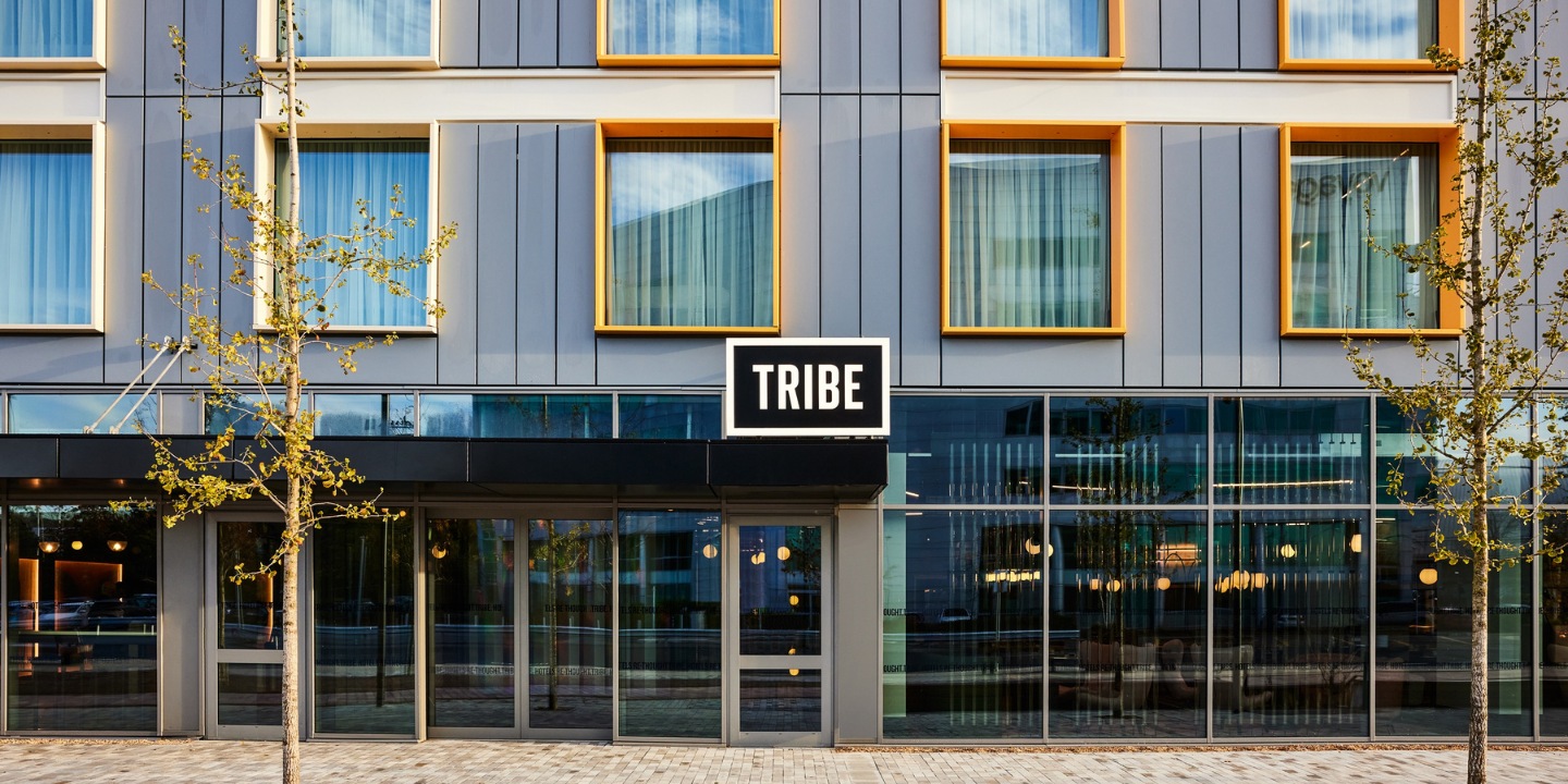 Modern hotel facade with large glass windows, a black "TRIBE" sign, and orange-accented window frames.