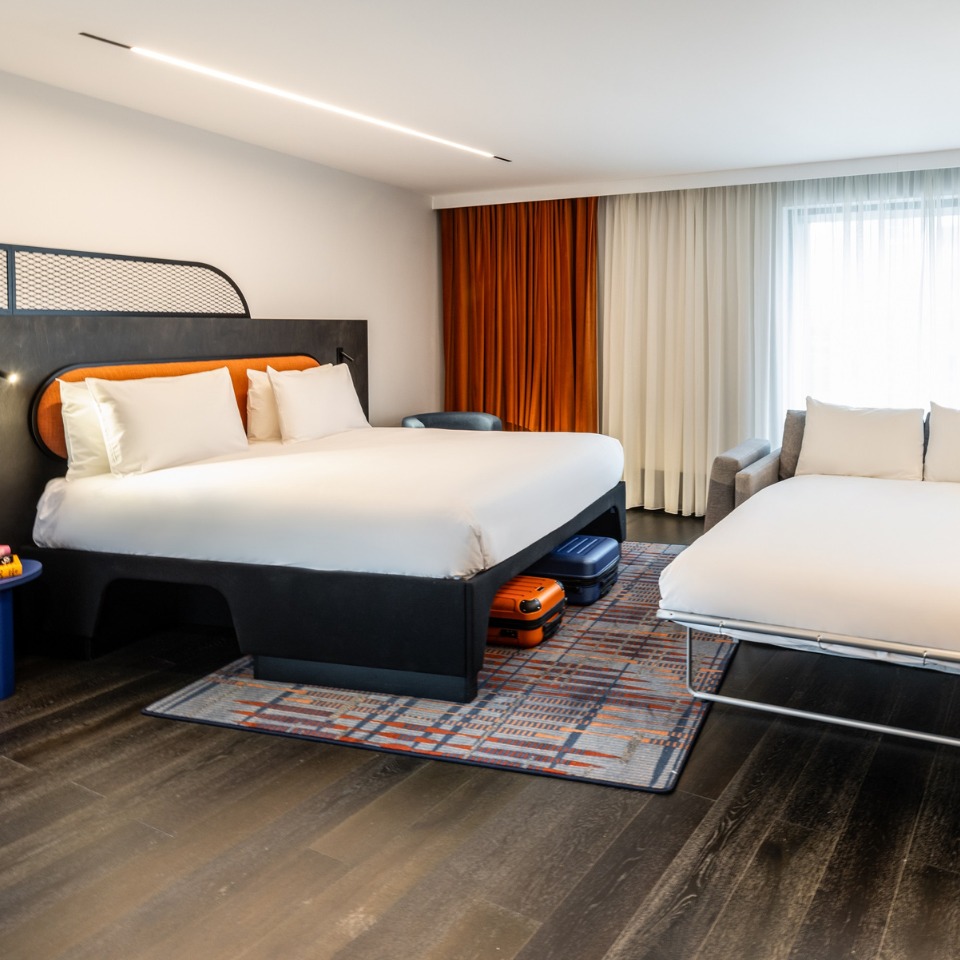 A modern hotel room featuring a king bed, a fold-out sofa, and stylish decor, with large windows and vibrant curtains.