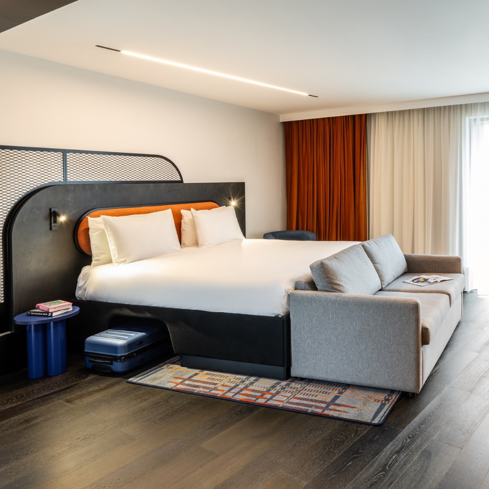 Hotel room featuring a king-sized bed, gray sofa, and stylish decor with orange accents and warm lighting.