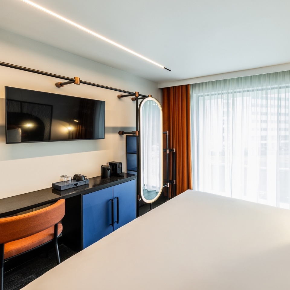 Modern hotel room featuring a large TV, elegant mirror, a coffee station, and a stylish orange chair by a bright window.