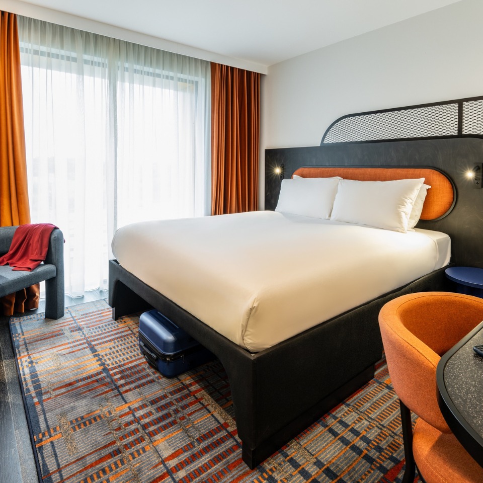 Hotel room featuring a large bed, bright curtains, a cozy sitting area, and stylish decor in orange and blue tones.